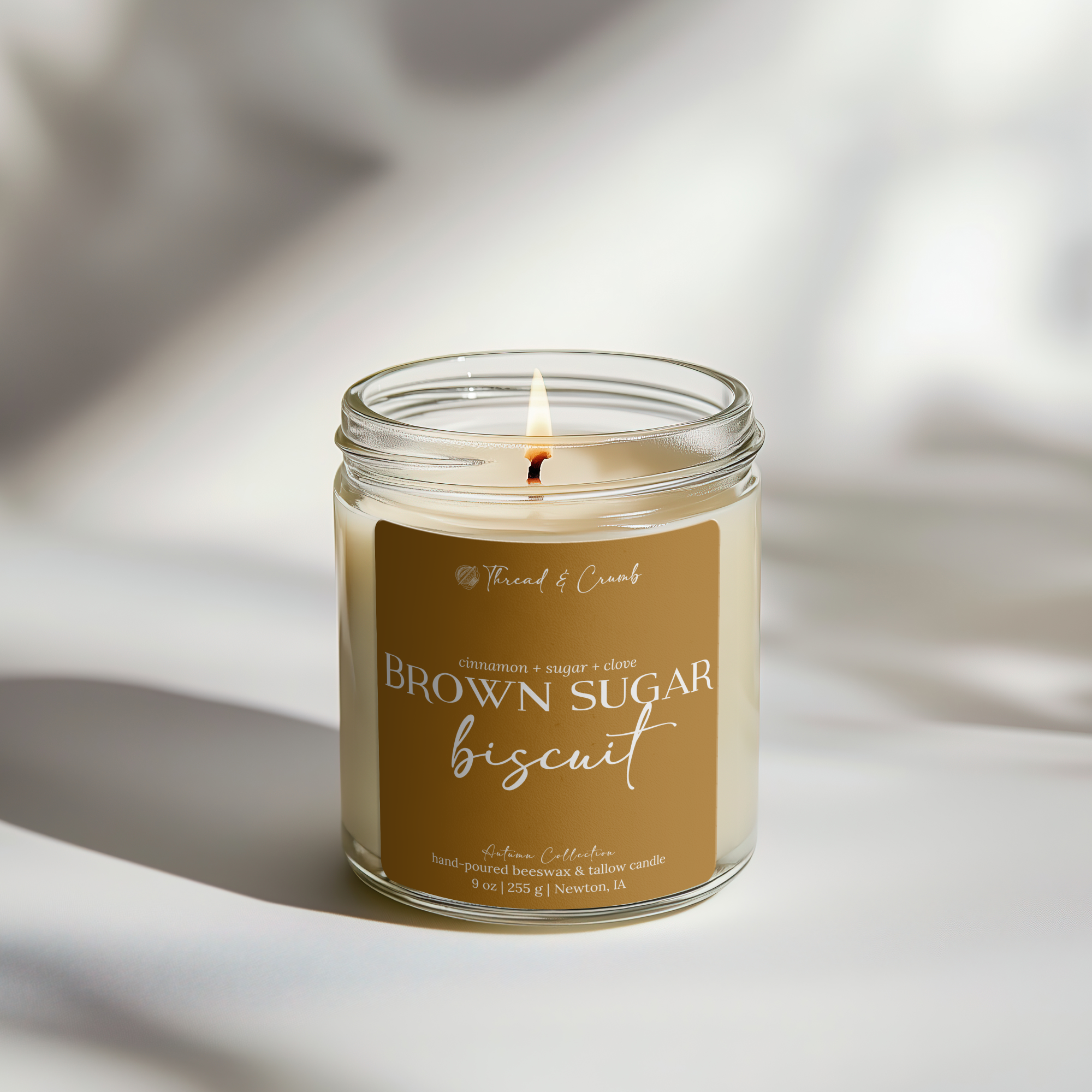 A lit jar candle labeled 'Brown Sugar Biscuit' by Thread & Crumb, set on a white surface with soft shadows and light.