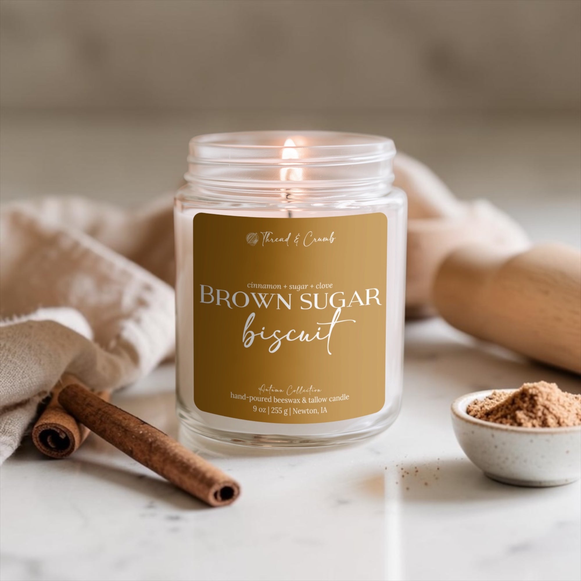 A lit candle in a clear glass jar with a tan label reading 'Brown Sugar Biscuit' by Thread & Crumb. The candle is styled on a marble surface with cinnamon sticks, a beige cloth, and a small bowl of brown powder in the background.