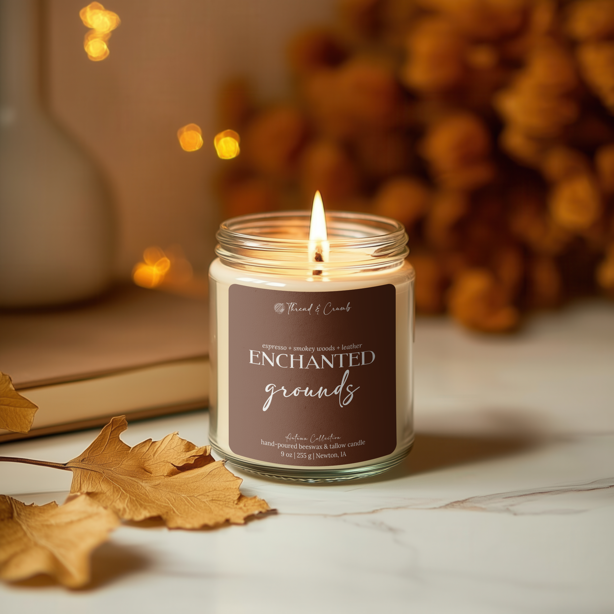 A lit candle in a clear glass jar sits on a light marble surface with autumn leaves and warm blurred decor in the background. The label reads 'Thread & Crumb' and 'Enchanted Grounds'.