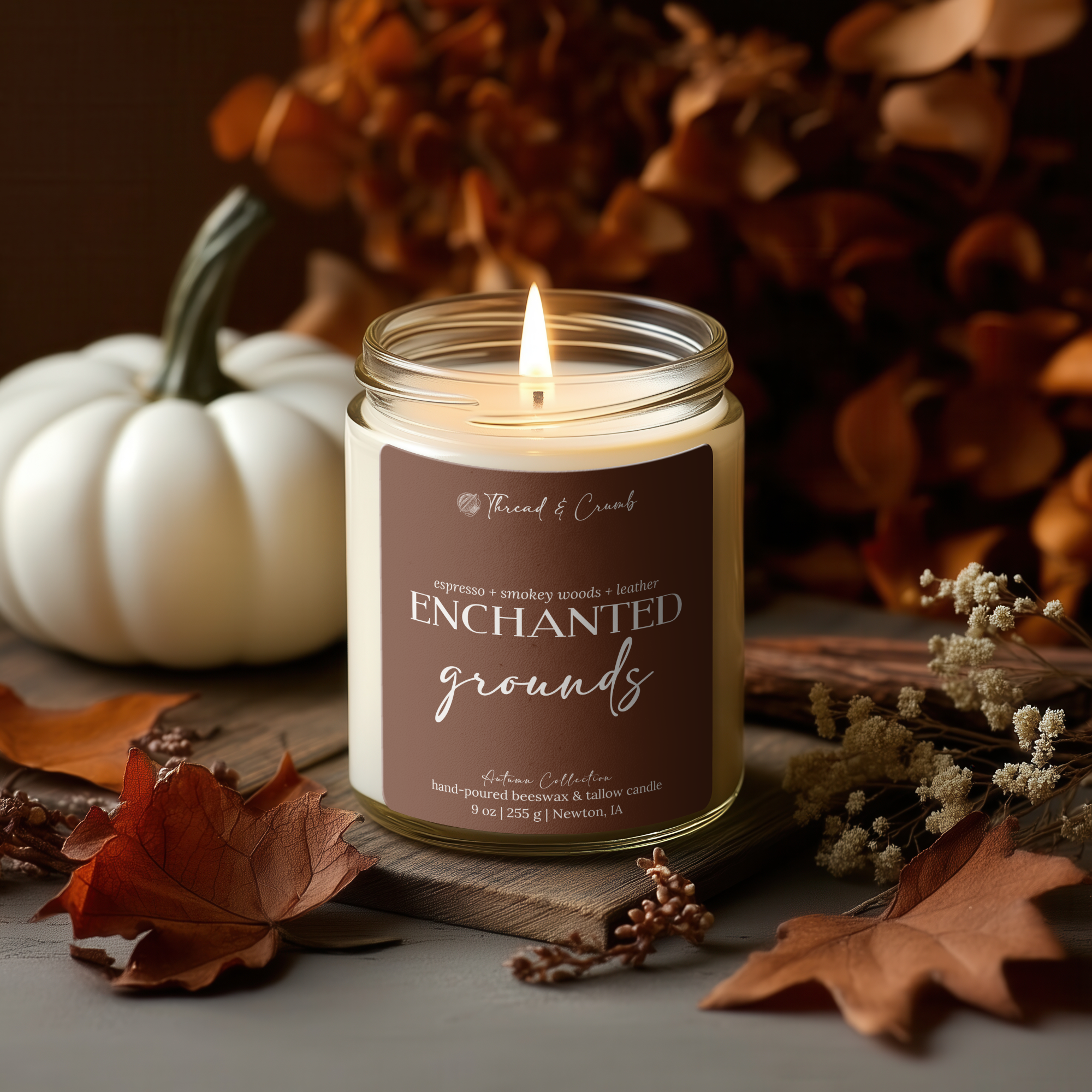 'Thread & Crumb' branded Enchanted Grounds fall coffee candle sitting on a wooden surface in a styled autumn scene styled with dried autumn leaves, a white pumpkin, and brown floral decor.