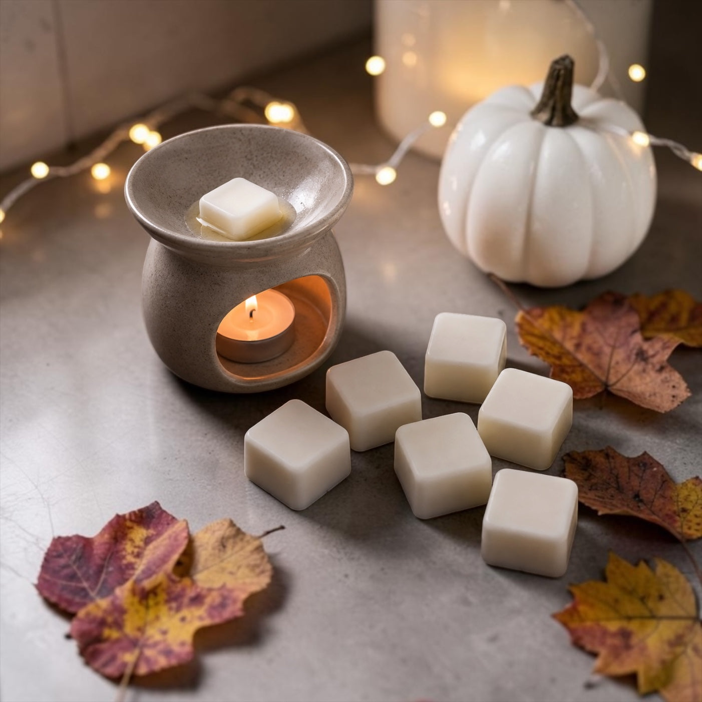 A set of white wax melts arranged on a gray surface next to a lit ceramic candle warmer and a white decorative pumpkin, with autumn leaves and warm fairy lights in the background.
