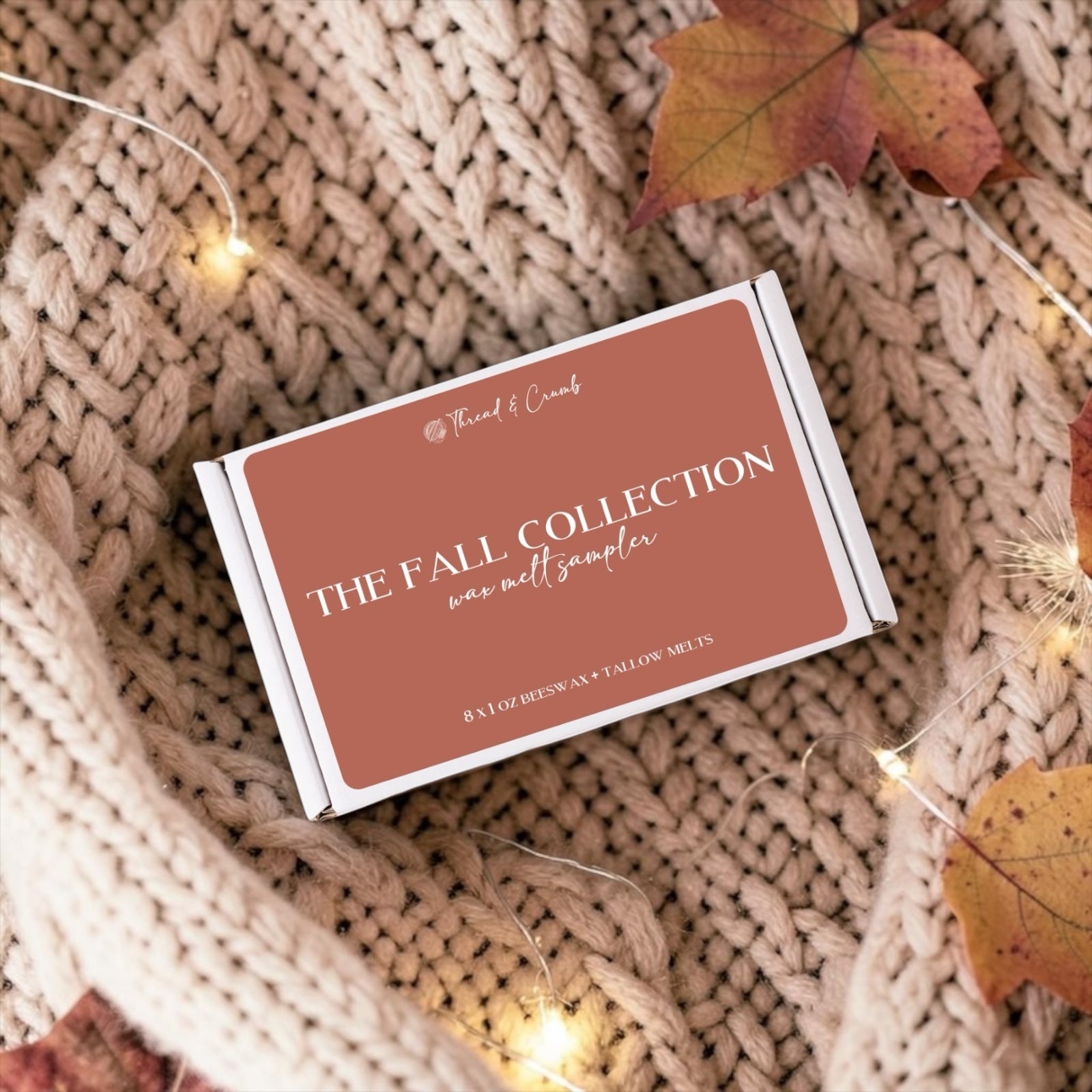 A rectangular product box labeled 'The Fall Collection wax melt sampler' by Thread & Crumb, placed on a beige chunky knit blanket with scattered autumn leaves and warm fairy lights.