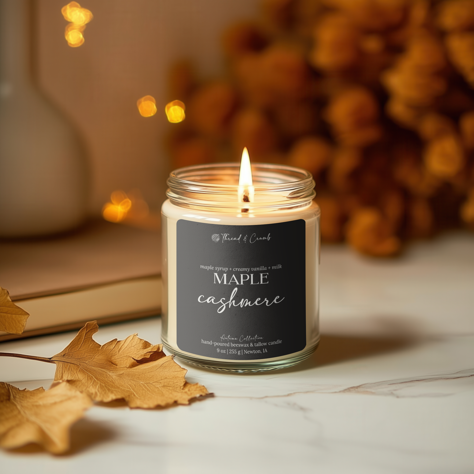 A lit jar candle labeled 'Maple Cashmere' by Thread & Crumb, set on a marble surface with autumn leaves and warm blurred background decor.