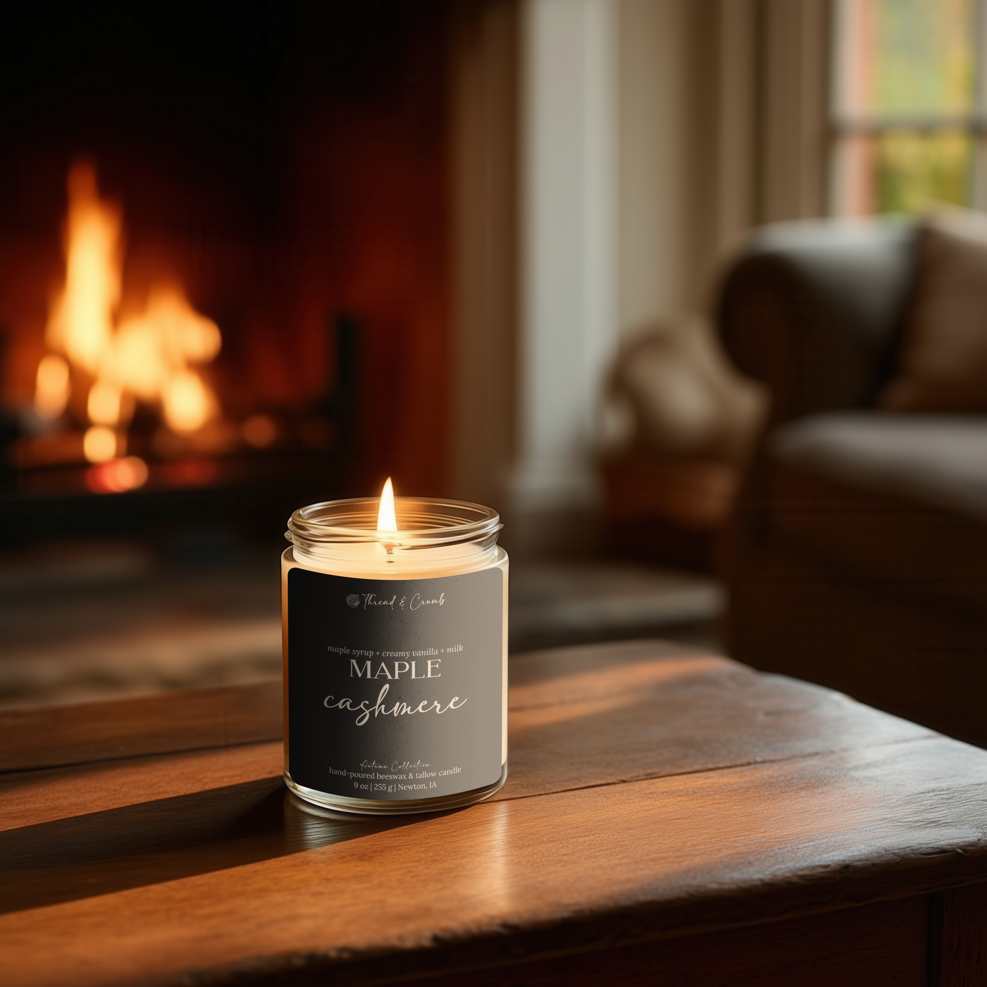 A lit 'Thread & Crumb' branded candle in a clear glass jar sits on a wooden table in a cozy living room with a blurred fireplace and sofa in the background. The label reads 'Maple Cashmere'.