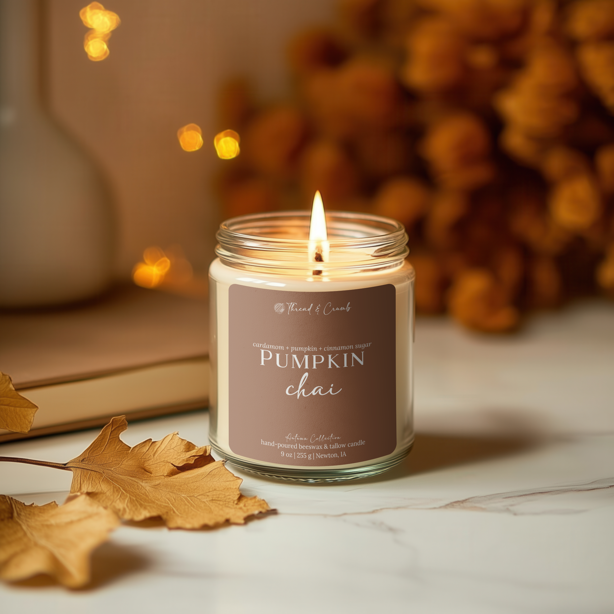 A lit Thread & Crumb branded Pumpkin Chai candle in a clear glass jar sits on a white marble surface with autumn leaves and warm blurred decor in the background.