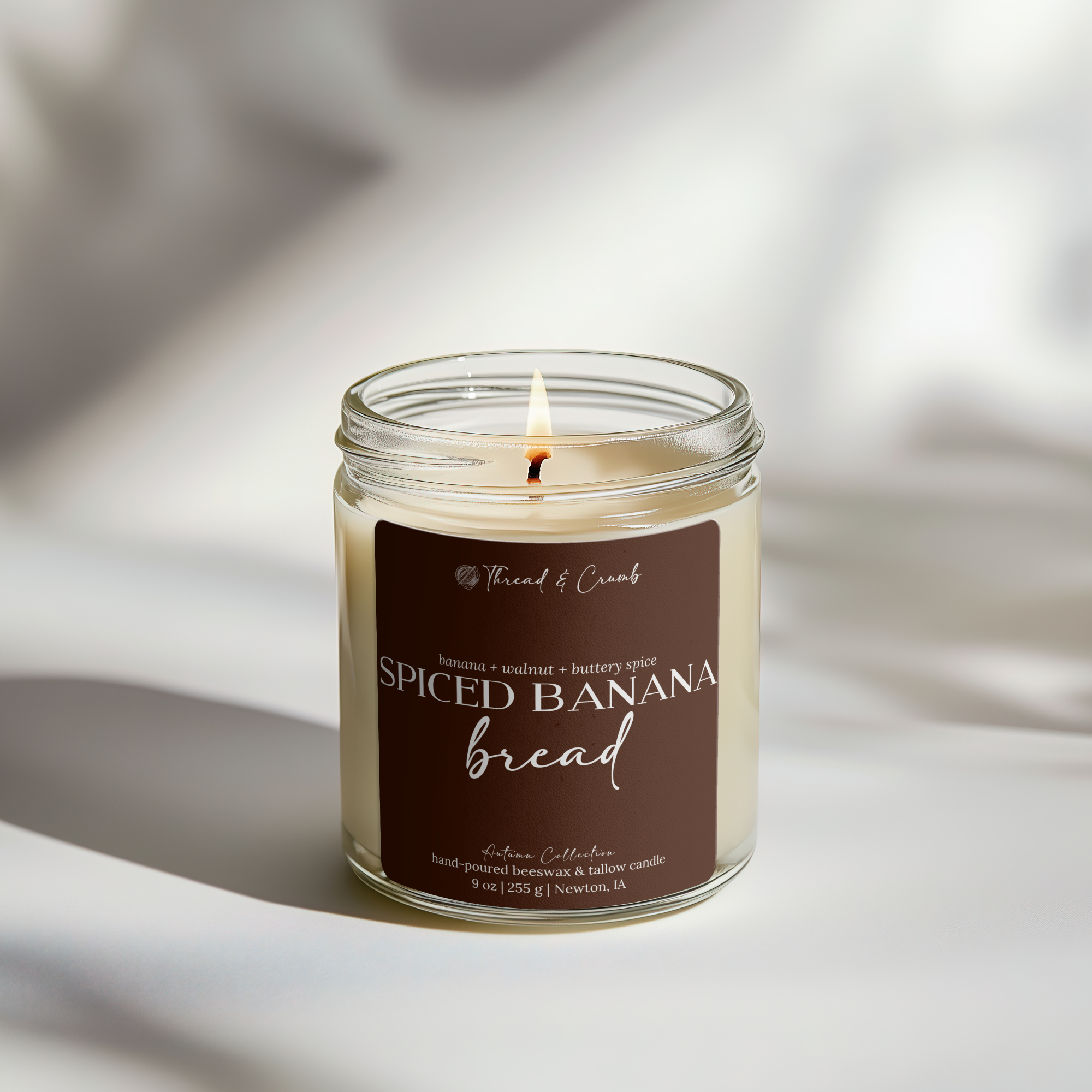 A lit candle in a clear glass jar with a brown label reading 'Thread & Crumb Spiced Banana Bread,' set on a softly lit white surface with warm shadows.