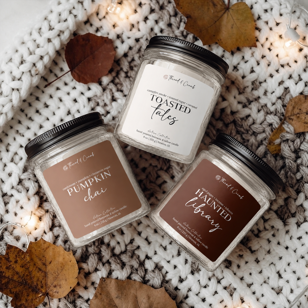 Three glass jar candles with black lids are arranged on a chunky knit blanket with scattered autumn leaves and warm string lights. The labels read 'Pumpkin Chai,' 'Toasted Tales,' and 'Haunted Library,' with the brand 'Thread & Crumb' visible on each label.