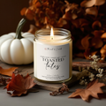A lit candle in a clear glass jar labeled 'Toasted Tales' by Thread & Crumb, styled on a wooden board with autumn leaves, dried flowers, a white pumpkin, and a warm brown background.